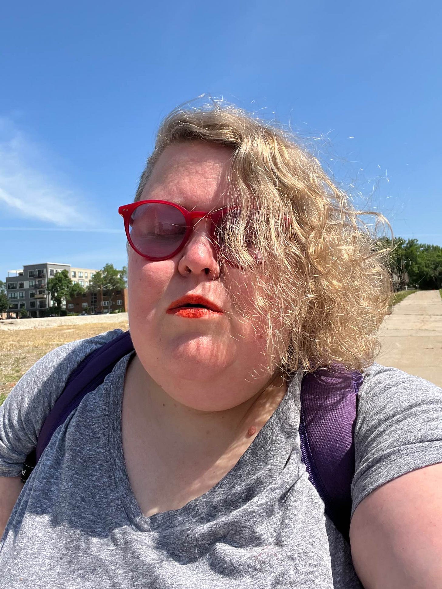 Me, Alison, a fat, white woman with asymmetrical curly blonde hair, red migraine glasses, and my eyes closed. Green trees in distance.