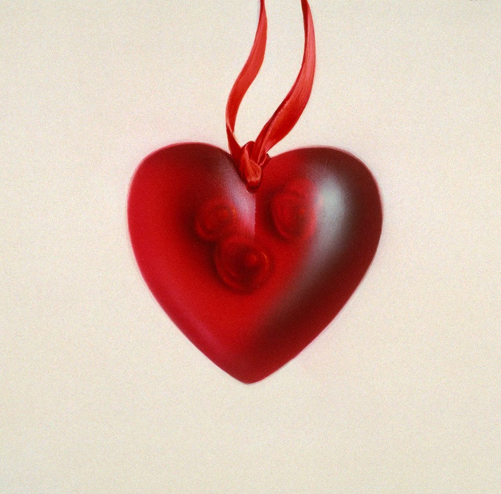 A transparent red glass heart hangs from a red ribbon against a neutral cream background. Inside four irregular shapes reminiscent of pearls sitting on clam shells overlap represent four chambers of the heart. Two of the shapes overlap almost completely while the other two only touch.