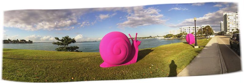 A large, bright-pink snail sculpture made of foam or something like it, placed on grass along a river bank. Above: clouds and a blue sky.