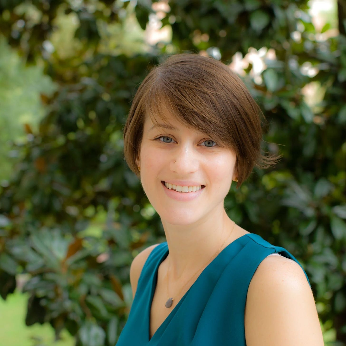 A photo of a smiling white woman with mid-length brown hair. She is wearing a teal, sleeveless shirt.