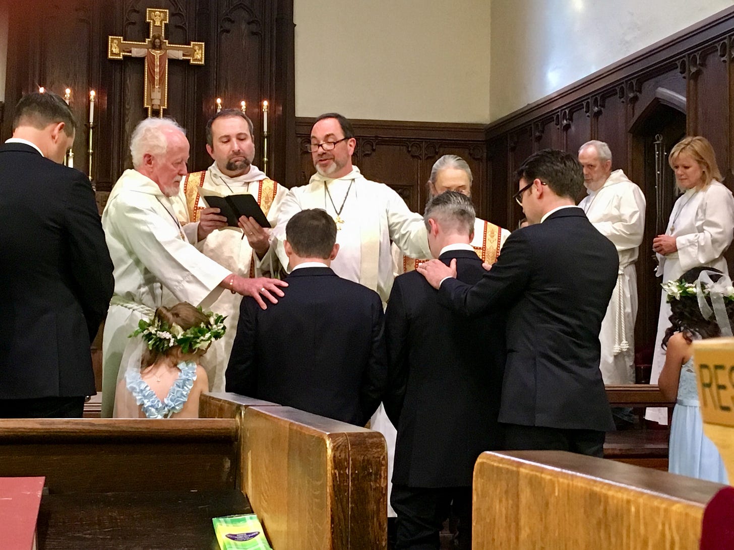 Priests, altar party, and wedding party gather in church as grooms kneel for blessing