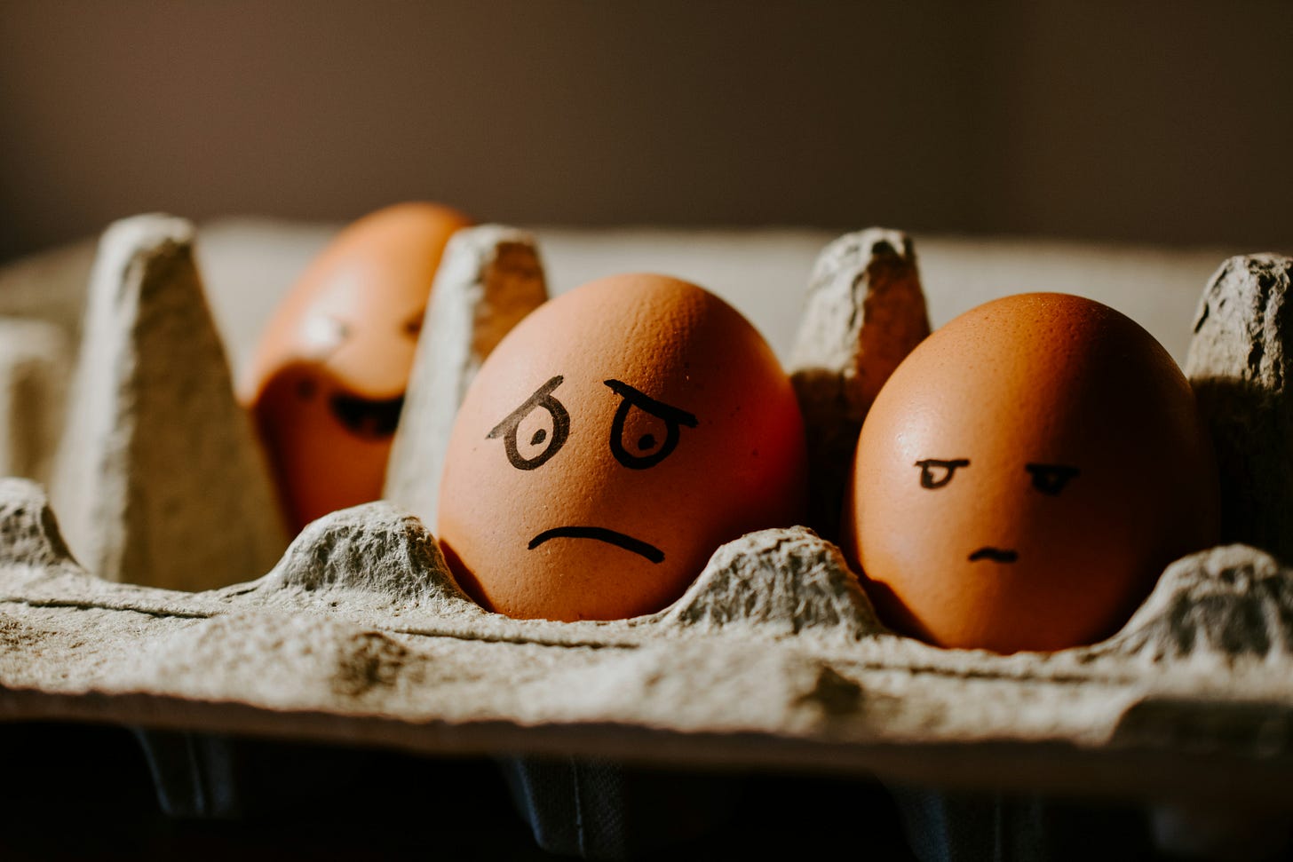 Photo of a grey cardboard egg box containing three eggs with faces drawn on them: one is laughing, another is looking suspiciously at the third, who is looking sad and confused. Photo of a grey cardboard egg box containing three eggs with faces drawn on them: one is laughing, another is looking suspiciously at the third, who is looking sad and confused.