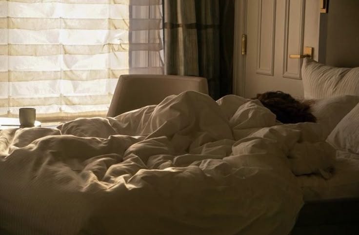 This may contain: an unmade bed with white sheets and pillows in front of a window at night