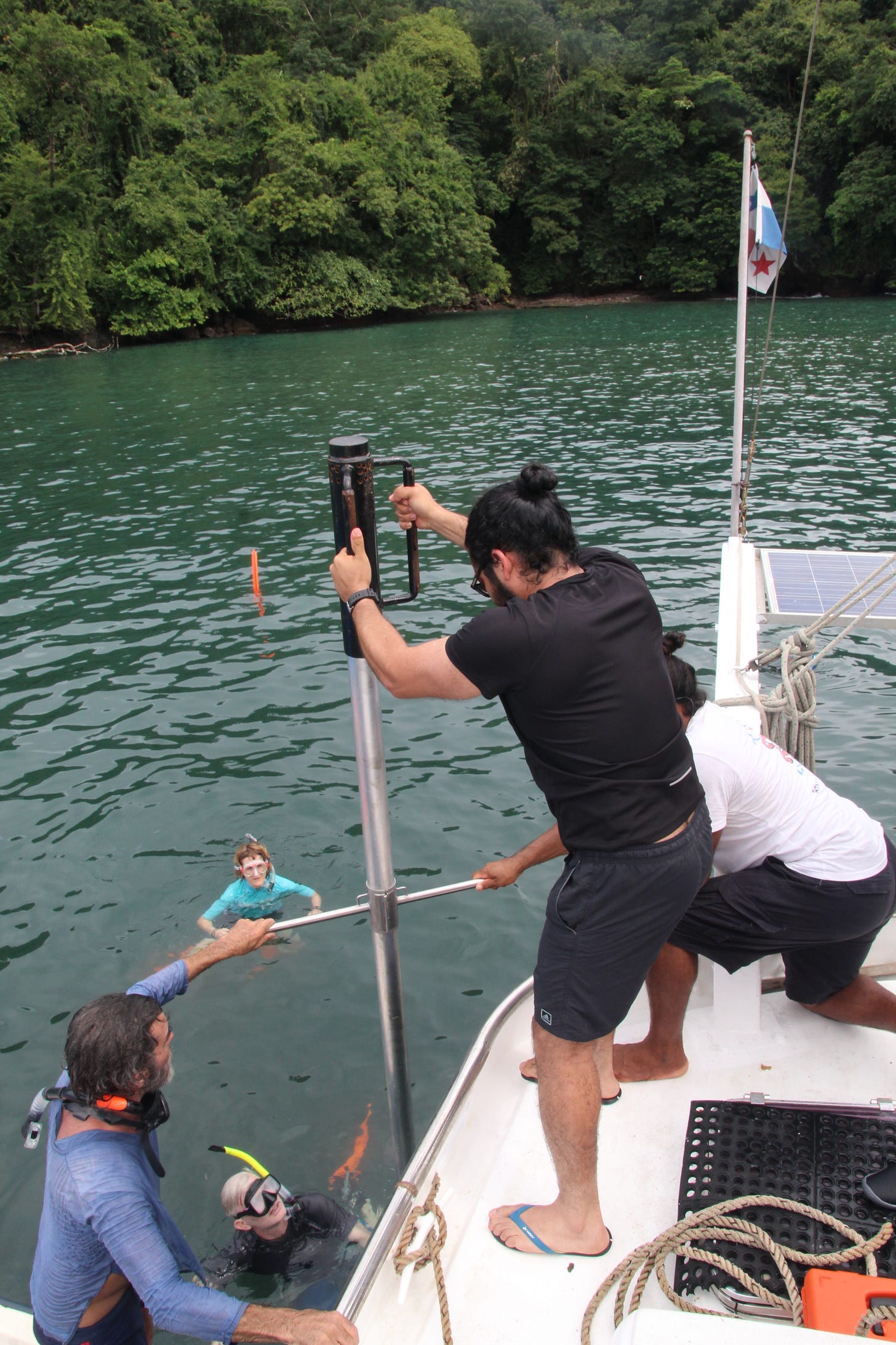 A lush green jungle frames the top of the photo, the ocean reflecting the greeness. On the lower edge, a team of scientists are hammeing a 10-meter long aluminium pipe into the ocean. Two men stand on the deck of a sail boat, one kneeling and holding the pipe in place, the other standing, employing force on the specially-designed, cylindrical hammer. Two people are in the water wearing snorkels. A fifth man is holding on to the sailboat and standing on an unseen dingy, helping hold the metal pipe.