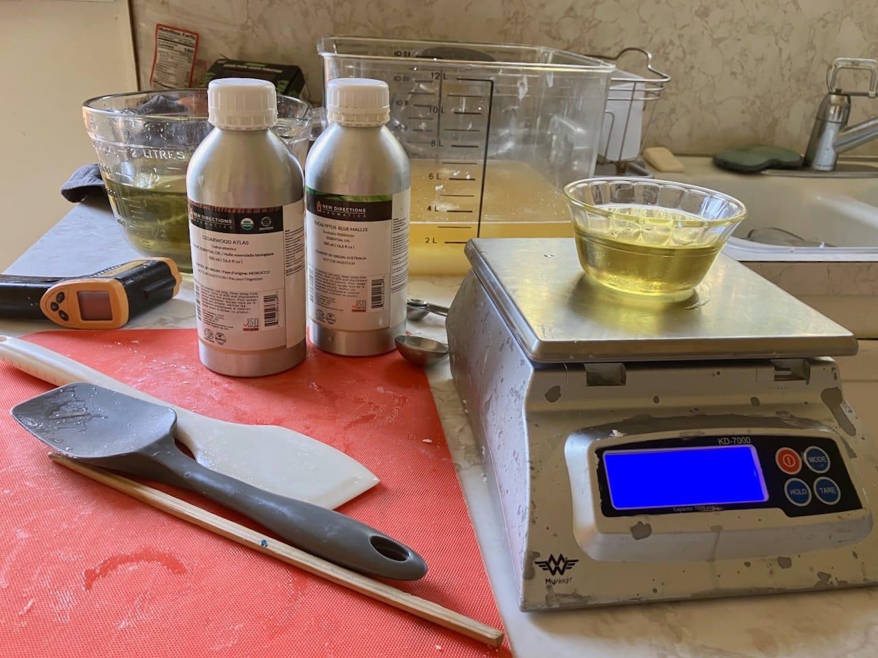 A kitchen workspace with essential oil bottles, a digital scale, and various tools set up for homemade soap making.