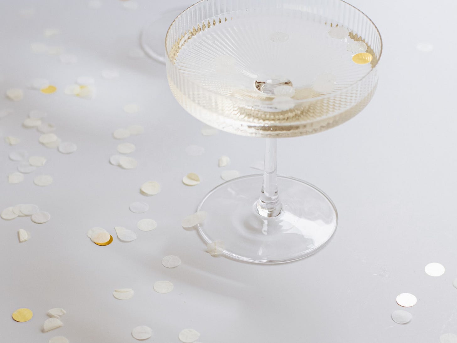 Glass of champagne surrounded by scattered white and gold confetti on a soft white surface, evoking a quiet moment of celebration or transition.
