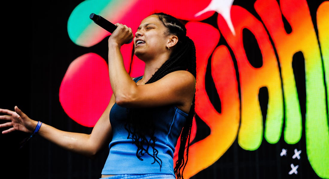 A woman with dark hair and a blue top holding a microphone, with a colourful background behind her A woman with dark hair and a blue top holding a microphone, with a colourful background behind her
