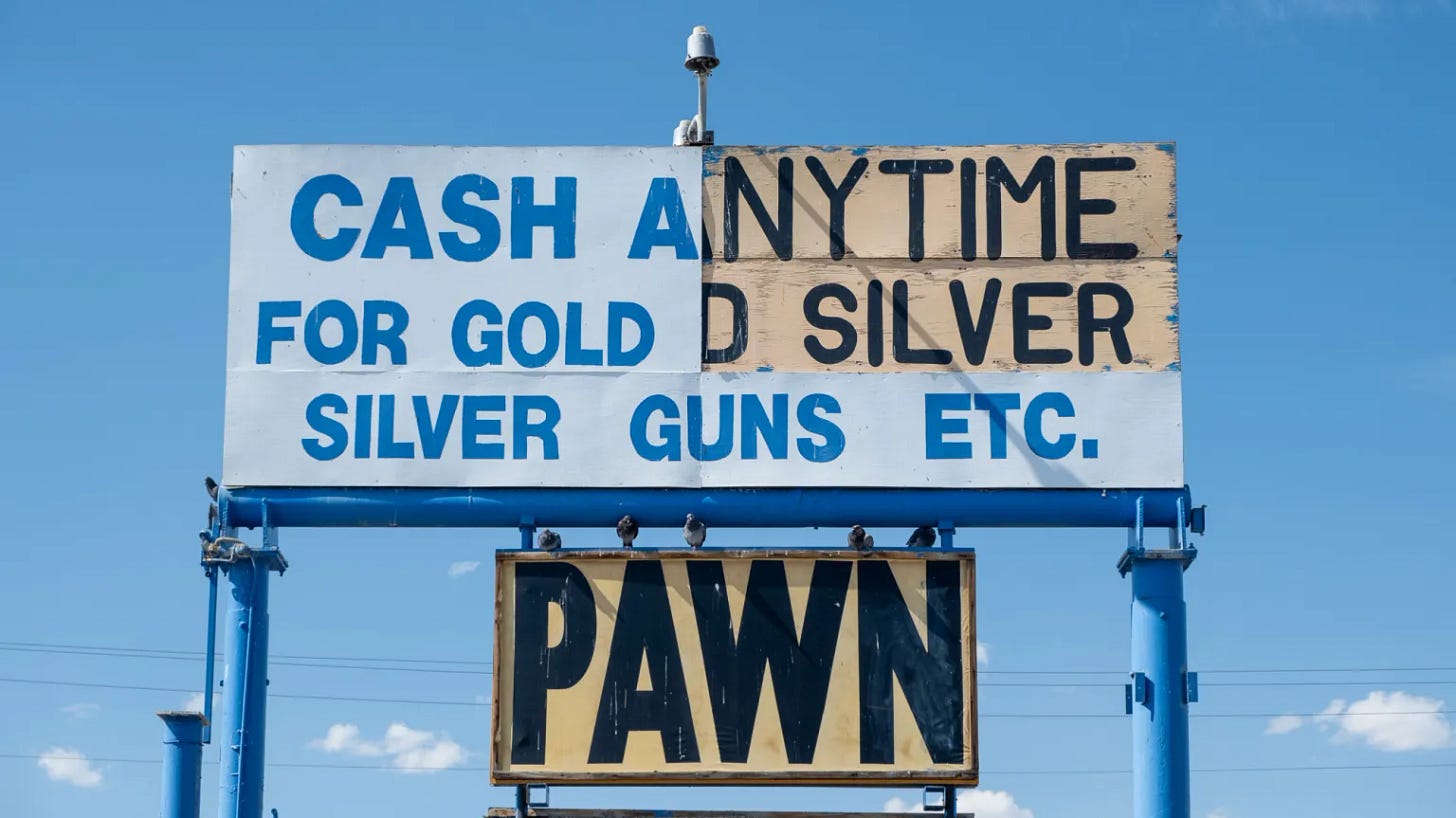 A large sign is shown against the backdrop of a bright blue sky. It is cobbled together from various pieces of painted wood and perched on tall blue metal supports. It reads in blue and black block text, "CASH ANY TIME FOR GOLD SILVER SILVER GUNS ETC. PAWN" A large sign is shown against the backdrop of a bright blue sky. It is cobbled together from various pieces of painted wood and perched on tall blue metal supports. It reads in blue and black block text, "CASH ANY TIME FOR GOLD SILVER SILVER GUNS ETC. PAWN"