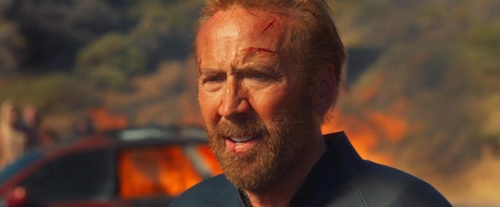 Nicolas Cage with Injuries Alt text: Nicolas Cage as the Surfer, his face bloodied and scratched, standing in front of a burning car in an intense moment from the film.