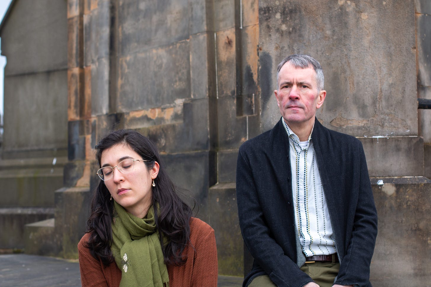 Portrait of Stephanie Lamprea and Alistair MacDonald, seated outside a cathedral wall. He is looking at the camera; she has her head tilted to one side, eyes closed as if dreaming.