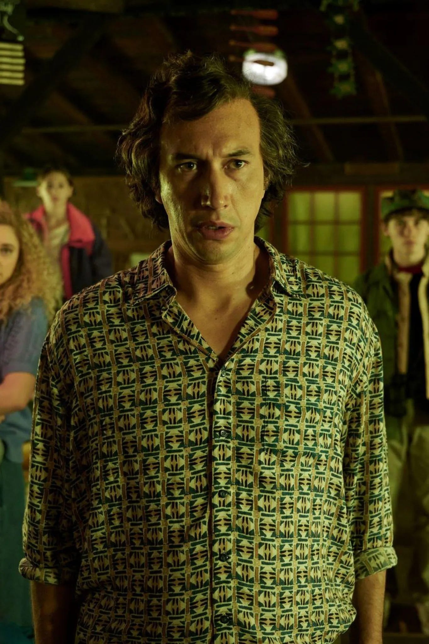 Adam Driver in White Noise. He has a receding hair line and wavy, gray, messy hair that is pushed back. It may be the hair, but his face appears to be slightly altered from his usual appearance using makeup or prosthetics.