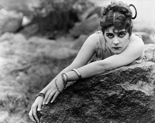 Publicity photo of Theda Bara with thick black eye makeup and many dangling braclets and earings. 