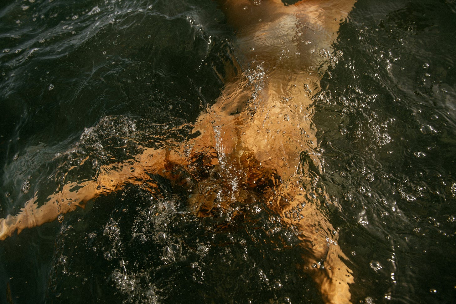 A body submerged just under dark water, lit gold, surrounded by bubbles.