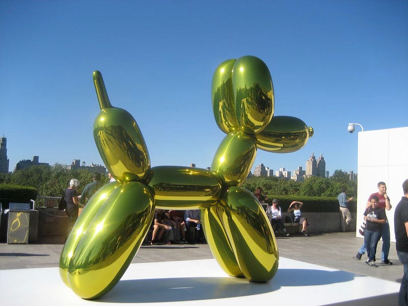 Koons Yellow Balloon Dog on the roof of a city building, with a blue sky behind it. A few people mingle around. 