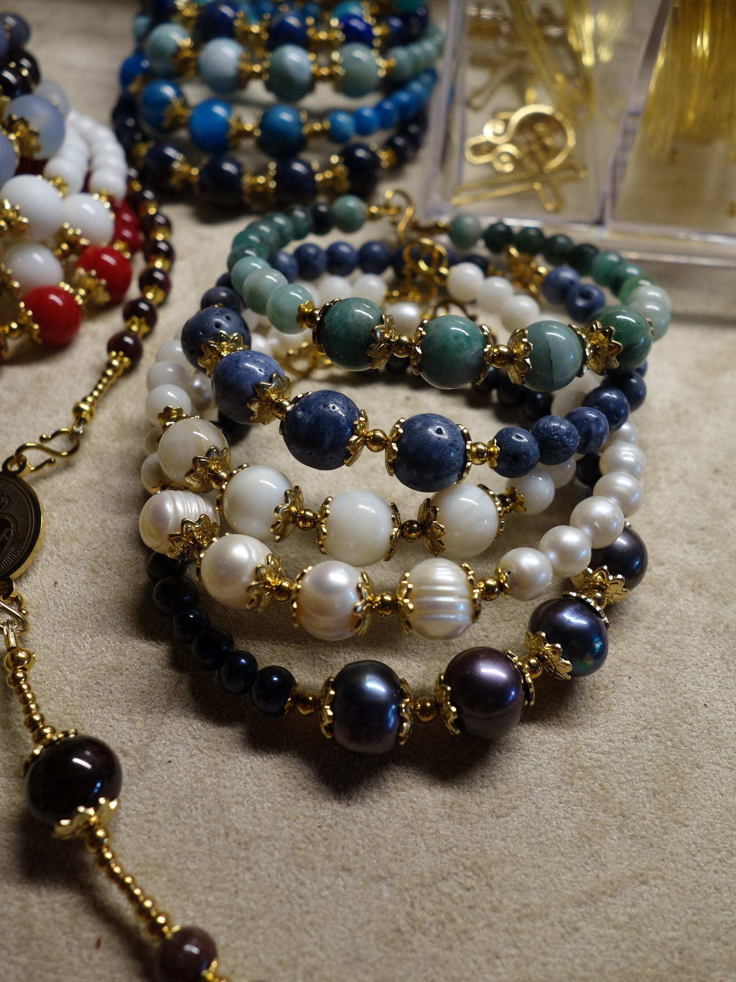 On the foreground: African Emerald, Blue Coral, White Throcus Shell aka Mother of Pearl, White Pearl (Cultivated Freshwater) Black Pearl (Cultivated Freshwater) - beads set on stainless steel memory wire with elaborate hooks. On the background: Lapis Lazuli, Larimar, Apatite, Natural Sapphire; 