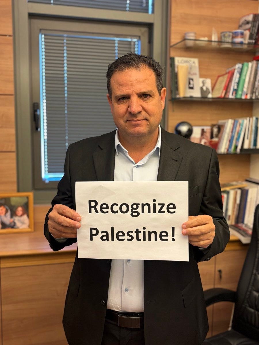 A man in a dark suit and light blue shirt stands in a wooden office with bookshelves filled with books and a framed photo, holding a white sign that reads Recognize Palestine! in English, with a globe on the shelf behind him and window blinds in the background.
