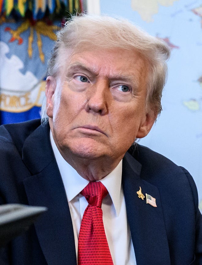 Trump against blue background with blue jacket, flag pin, stupid red tie; his head is cocked and he looks distracted and not magic