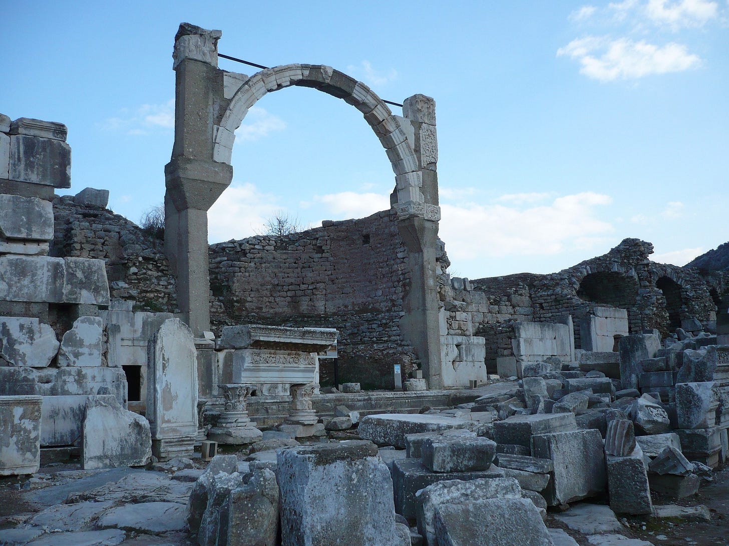 Blue sky. Ruins in lower light of the sun behind them. An arch supported by a metal cross-brace overlooking large fallen stones. A wall of smaller stones to the side and behind the arch. Looking towards but at a diagonal the arch.