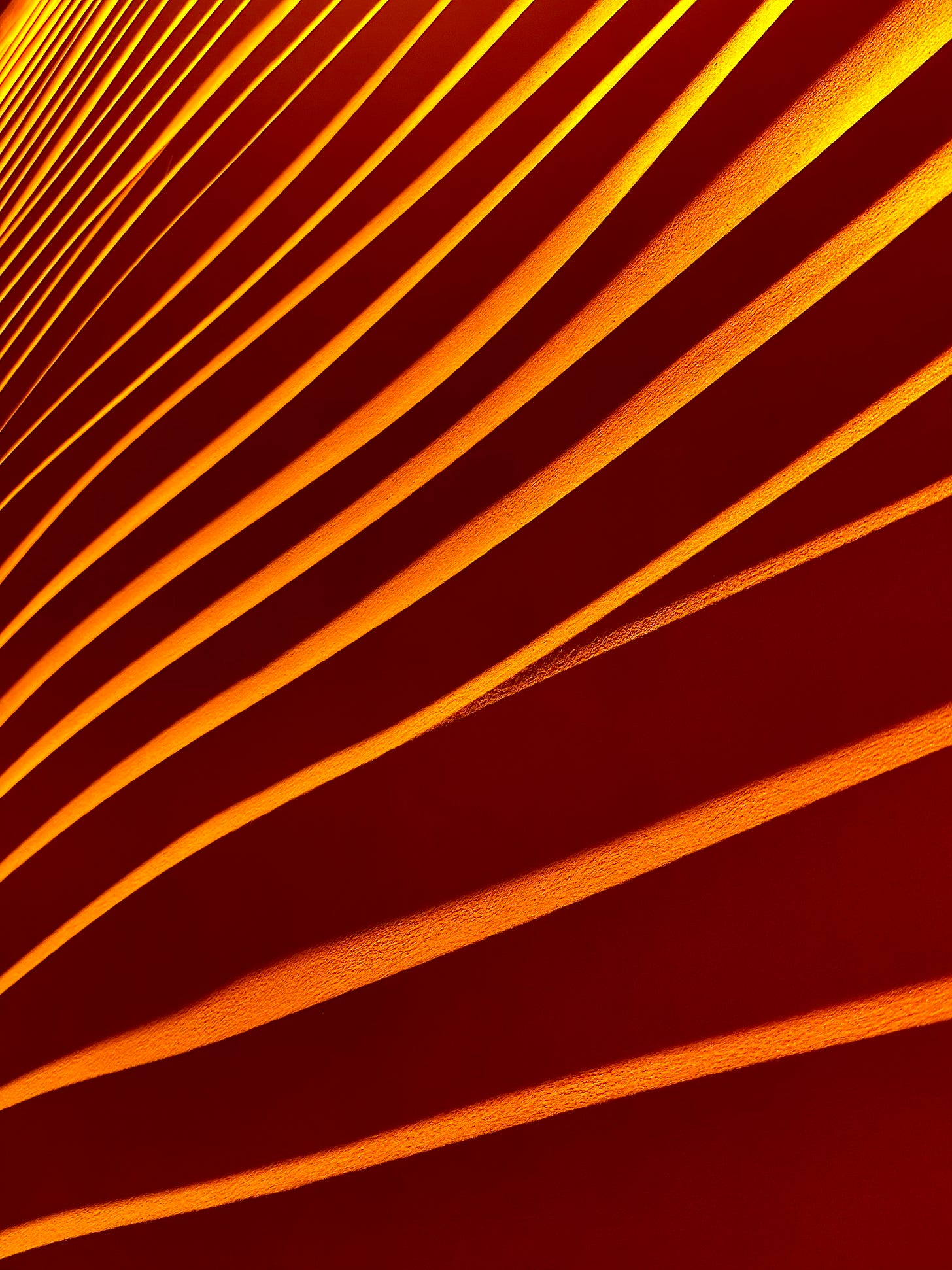 A close up photo of a textured wall with ridges running at diagonal angle. The light throws a deep orange colour onto the upper sides of the ridges which contrasts dramatically with the shadowy undersides.