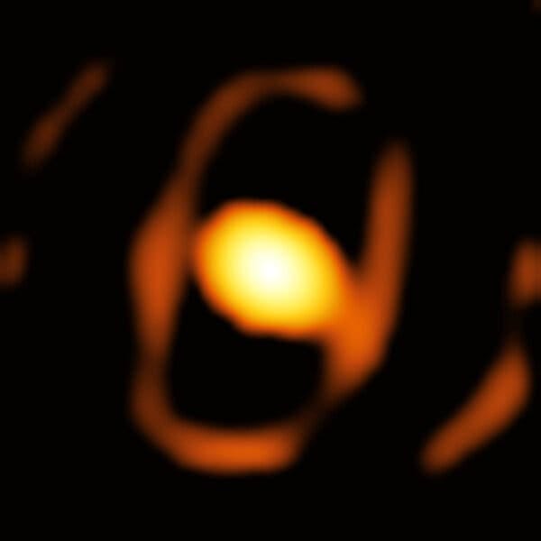 A bright orange-yellow dot surrounded by rings of orange material, out of focus, against the blackness of space.