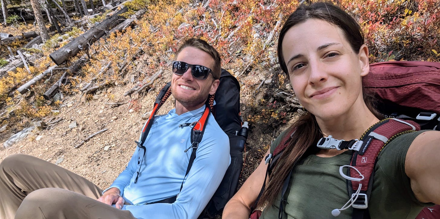 Two hikers taking a break on a mountain trail, sitting on the ground with backpacks resting against their backs. The woman on the right smiles at the camera, while the man beside her, wearing sunglasses and a light blue shirt, also smiles. Surrounding them are dry, rocky terrain and patches of autumn foliage, creating a warm and relaxed outdoor scene.