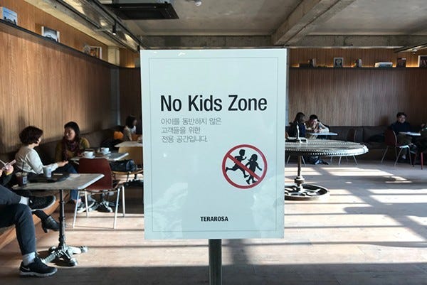 a room with tables and chairs with a sign that reads, "No Kids Zone" in English with Korean characters underneath and an illustration of kids running inside a red crossed out circle a room with tables and chairs with a sign that reads, "No Kids Zone" in English with Korean characters underneath and an illustration of kids running inside a red crossed out circle