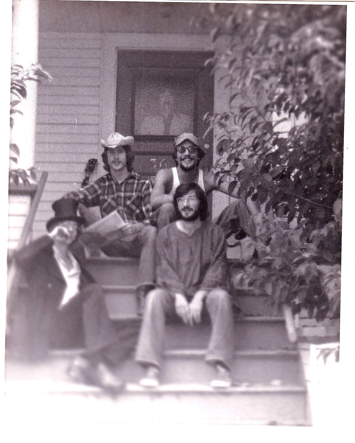 Four young men sitting on the steps outside a house and one standing behind a window in the door