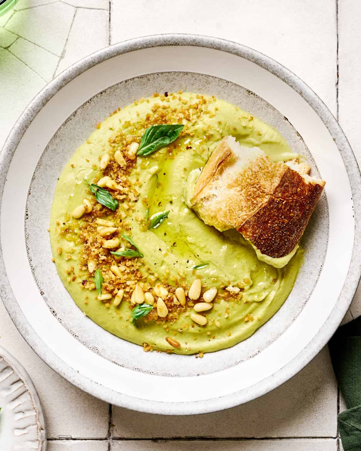 Hunk of sourdough in a bowl of creamy vegan broccoli soup in a white bowl on white tile table.