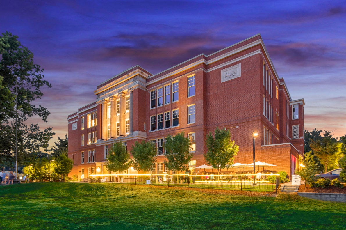 A brick building lit with warm lights at twilight, surrounded by trees and green lawn