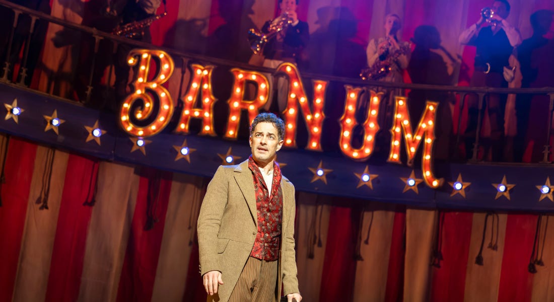 A man - Lee Mead - wearing a brown suit with red waistcoat. He is standing in front of a red and white backdrop with a sign that says 'Barnum' behind him A man - Lee Mead - wearing a brown suit with red waistcoat. He is standing in front of a red and white backdrop with a sign that says 'Barnum' behind him