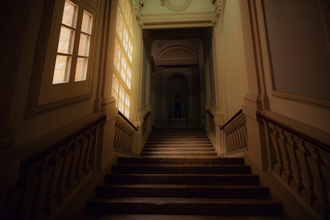 Dimly lit grand staircase with sunlight streaming through windows