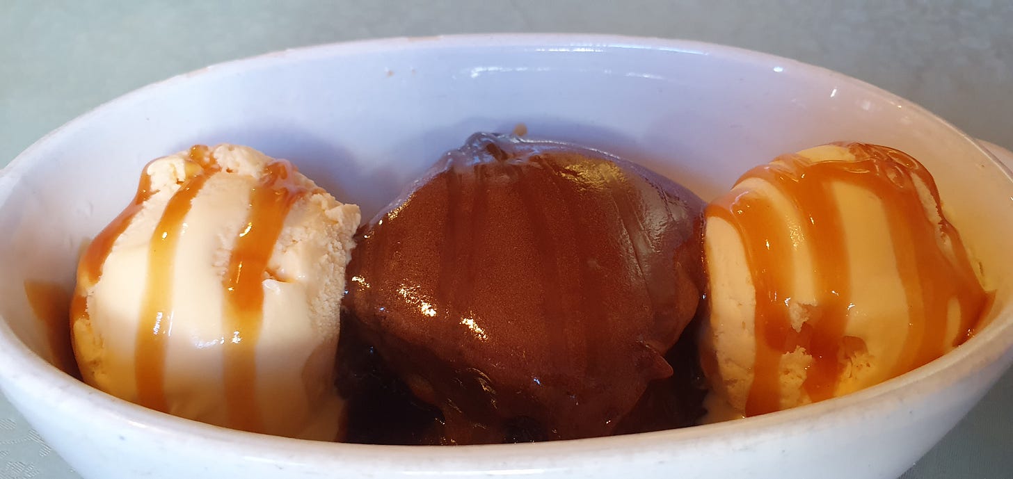 A serving of rich and sweet sticky toffee pudding is shown in a ceramic bowl, with two balls of vanilla ice cream.