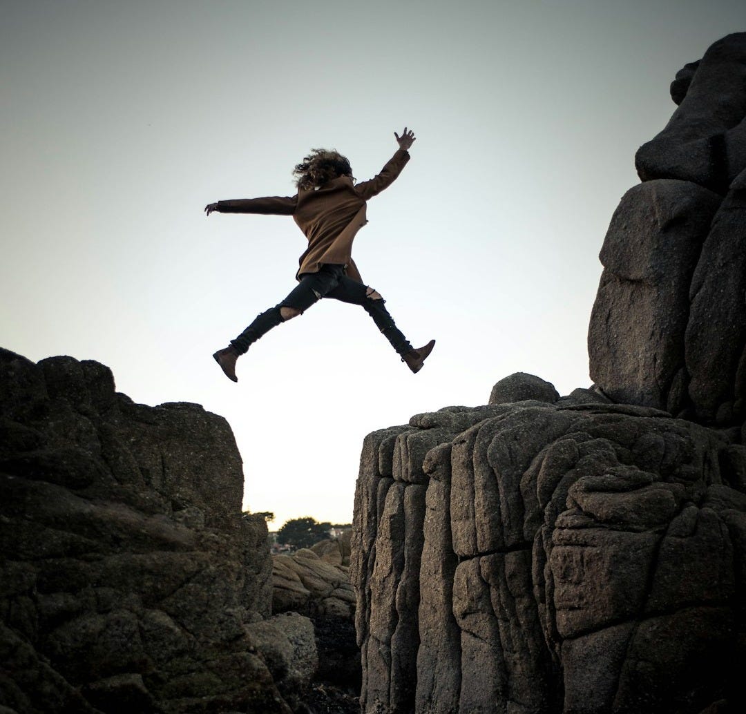 person jumping on big rock under gray and white sky during daytime