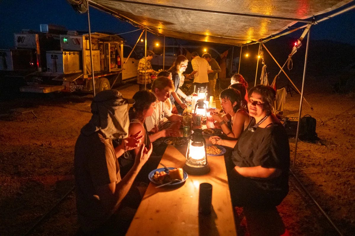 A group of people sit at a long table under a tarp, sharing a meal at night by lantern light. In the background another group of people huddle around the kitchen area preparing and serving food. The Land Arts truck can be seen in the background with doors open, revealing a pantry of cooking goods. A group of people sit at a long table under a tarp, sharing a meal at night by lantern light. In the background another group of people huddle around the kitchen area preparing and serving food. The Land Arts truck can be seen in the background with doors open, revealing a pantry of cooking goods.