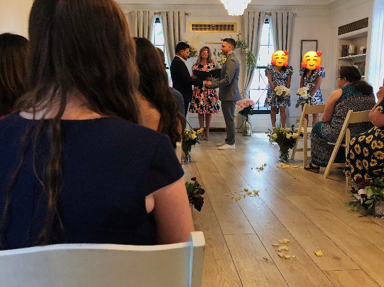 Two men in suits at the end of a cozy house-like aisle exchanging vows with friends and family seated around them.