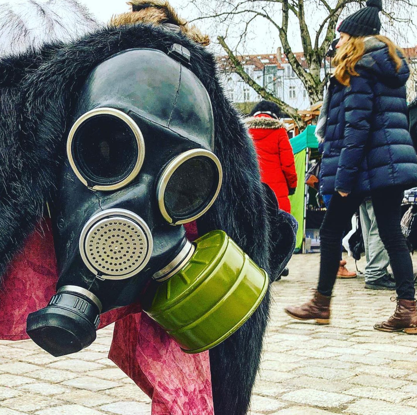 Close-up of a vintage gas mask with a green filter canister on display at a Berlin flea market, with people in winter coats walking past in the background.