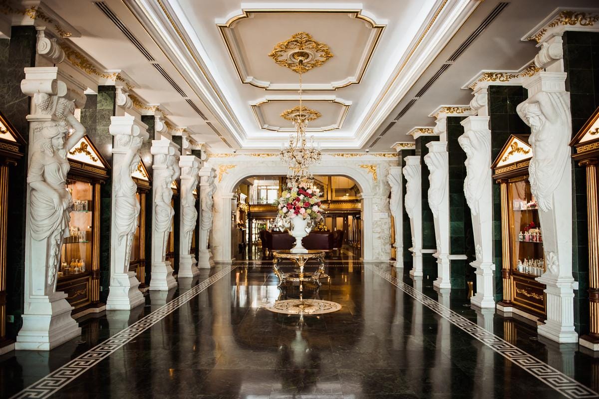 An ornate hotel corridor lined with marble caryatid columns sculpted in the form of draped female figures. The ceiling features intricate gold detailing, and a grand chandelier hangs over a central floral arrangement atop a gilded table.