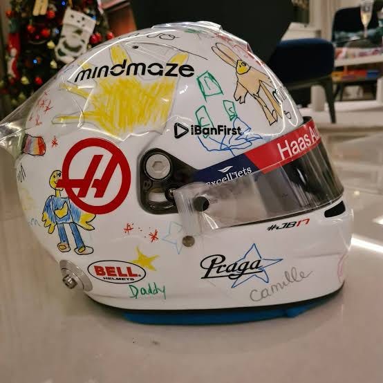 Romain Grosjean holding a white racing helmet with a tinted visor. The helmet features the Haas F1 logo, Bell branding, and various colorful drawings and signatures, including "mindmaze," "iBanFirst," "Praga," "Daddy," and "Camille." The background includes a modern indoor setting with a Christmas tree and posters of the Eiffel Tower.