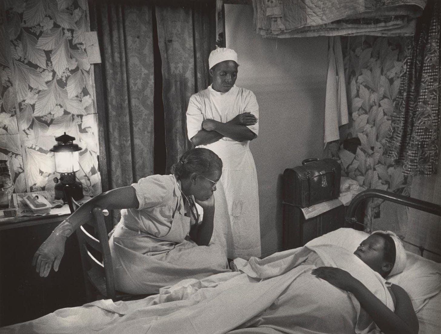 two Black woman, one in a cap and standing, tend to a woman, also wearing a cap, on a bed with blankets pulled to her chin