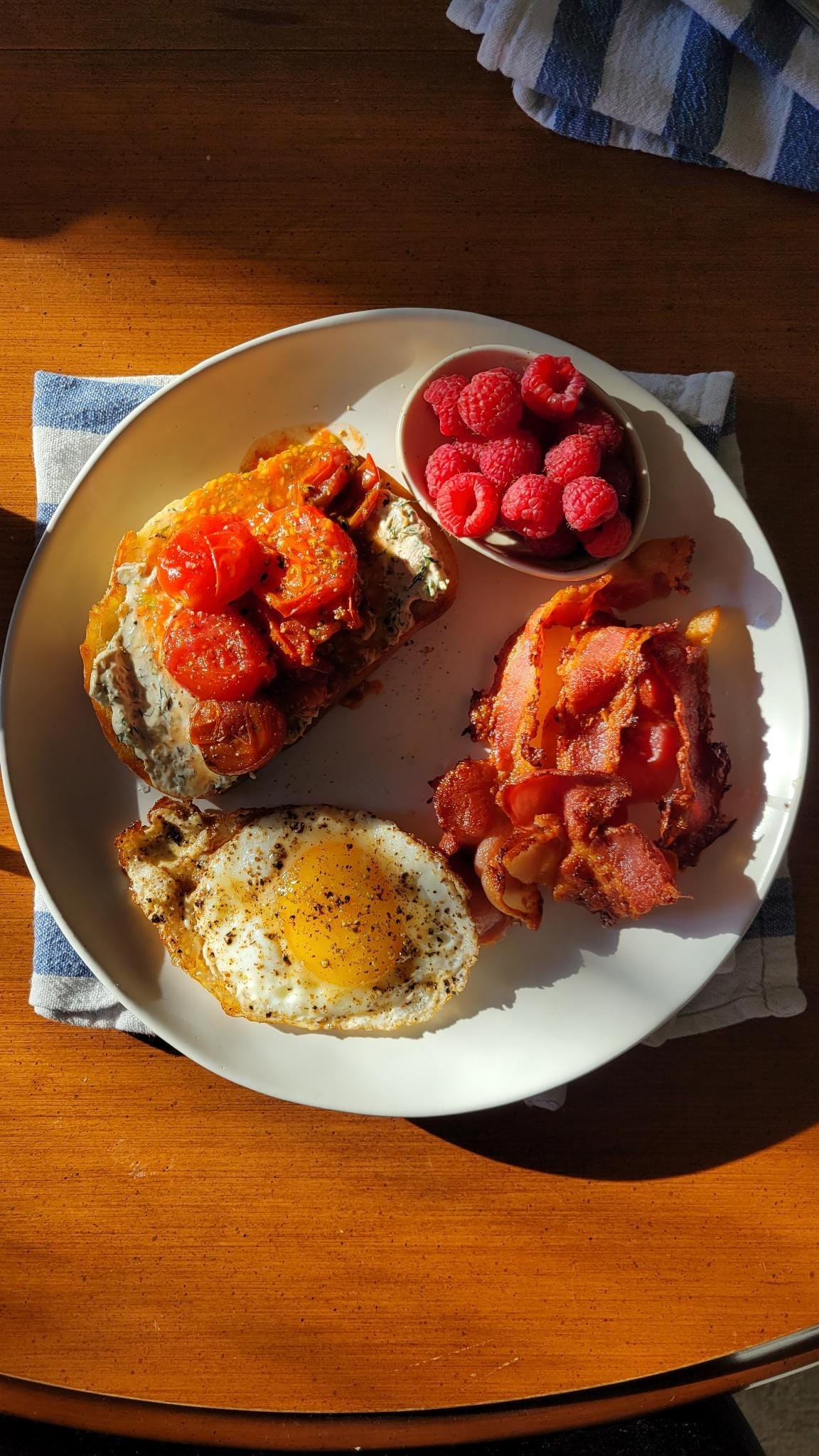 A white plate covered with a piece of stewed tomatoes on toast, an egg, a small pile of bacon and a small ceramic bowl full of raspberries. The sunlight hits it an illuminates it in great contrast to the wood table it sits on