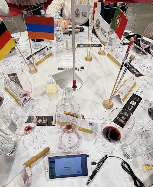 Close-up of the Concours Mondial de Bruxelles judging tablet displaying 'Congratulations! There is no sample left to be tasted,' with wine glasses, national flags, and a keyboard on the tasting table