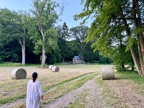 Golden evening light on the château grounds: a child in a white nightgown walking toward the coach house past hay bales; a girl on a stone path with the château behind; a man and two children pushing a large round hay bale.