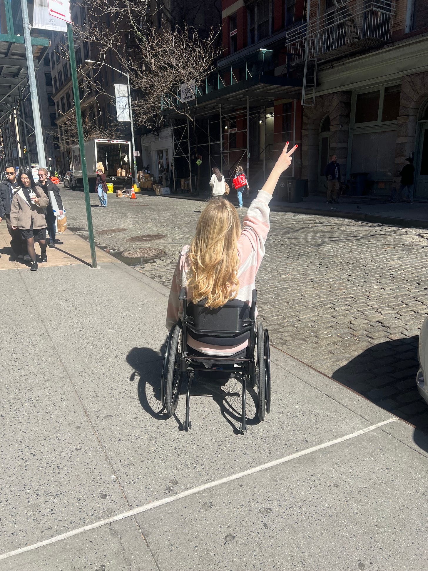 Jayne in a wheel chair facing the streets of NYC in making a peace sign. 