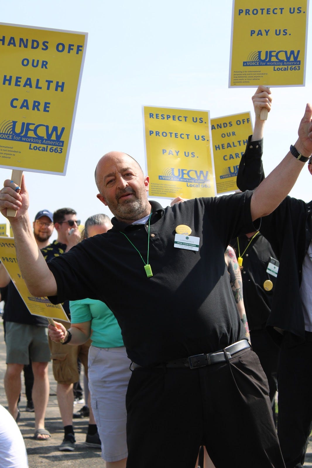 a white bald mine wearing a black polo shirt is protesting in a crowd and carrying a yellow sign with blue text that reads "hands off our health care UFCW local 663"