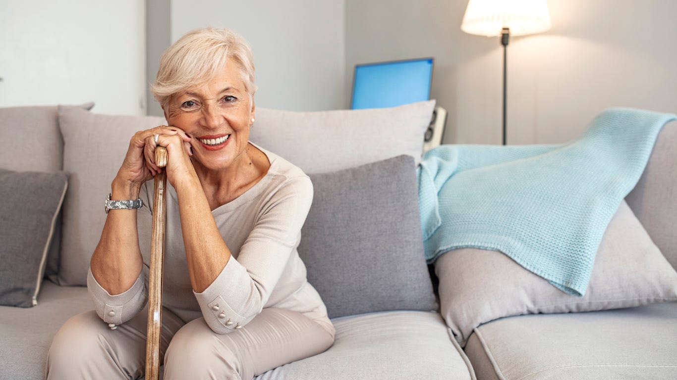 Smiling gray-haired white woman with cain in hand sitting on couch at home.