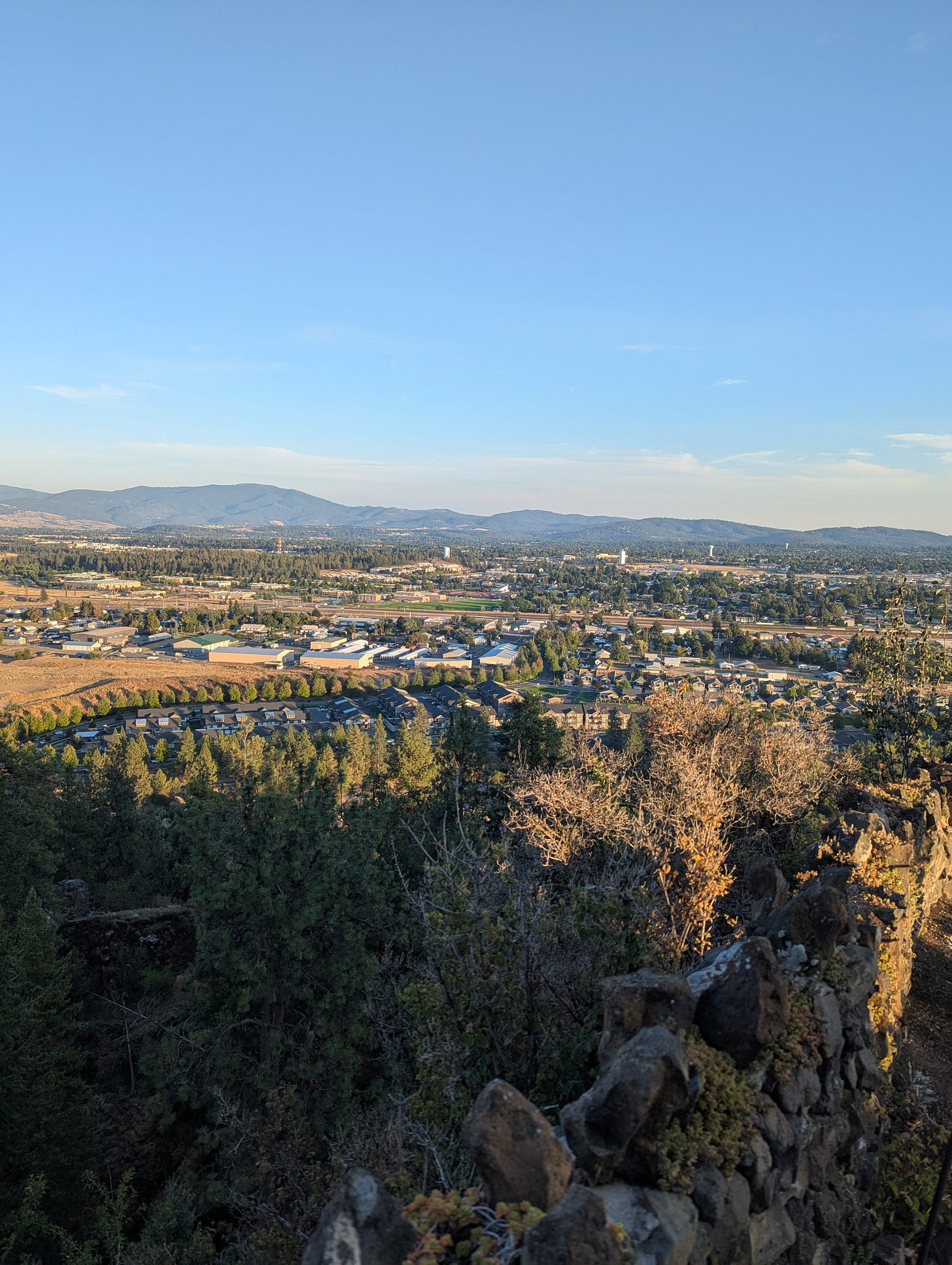 Picture of the sun setting over a valley of trees with mountains kissing a blue sky in the background. The light is golden hour light, barely touching the landscape below.