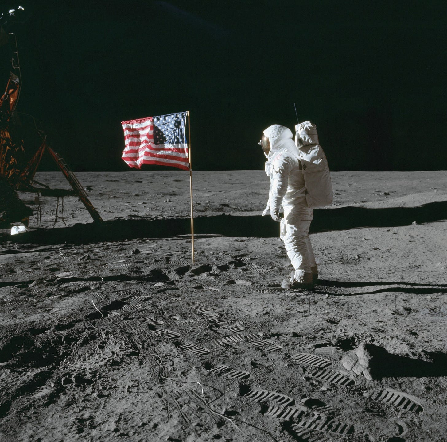 Astronaut Buzz Aldrin looks at a United States flag as he stands to the right of it. He is wearing a white spacesuit. The lunar module is on the left side of the flag. Bootprints in the gray lunar soil trail from the right in the foreground to the gold lunar module in the back. The darkness of space provides a contrasting backdrop to the image. The faint crosses on the image are markings that are used to calibrate distance and heights in photos taken on the lunar surface. Astronaut Buzz Aldrin looks at a United States flag as he stands to the right of it. He is wearing a white spacesuit. The lunar module is on the left side of the flag. Bootprints in the gray lunar soil trail from the right in the foreground to the gold lunar module in the back. The darkness of space provides a contrasting backdrop to the image. The faint crosses on the image are markings that are used to calibrate distance and heights in photos taken on the lunar surface.