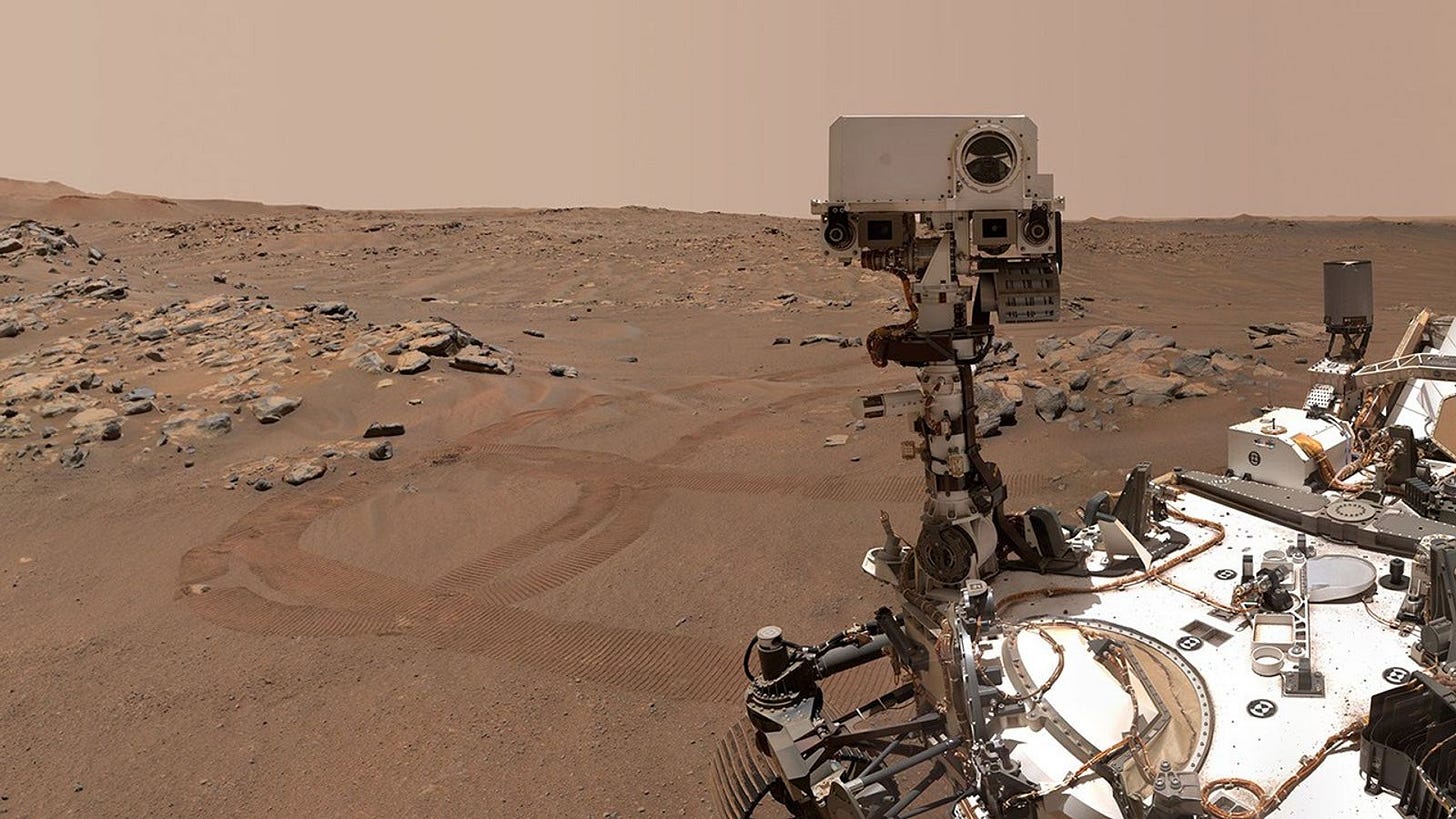 A selfie image of the Perseverance rover with flat Martian land in the background. The background is brown and dusty with some stray rocks and rover tracks in the soil, and the sky appears a pinkish beige. Credit: NASA/JPL-Caltech/MSSS