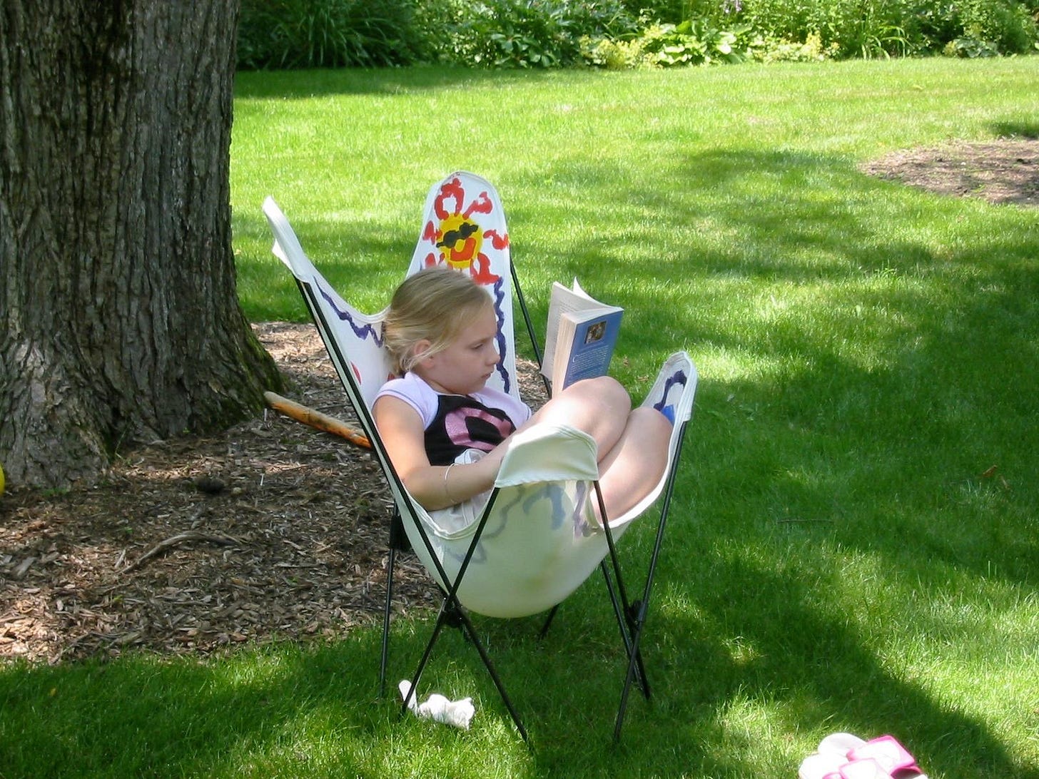 Young girl curled up in a hand-painted canvas butterfly chair, reading a book in a backyard under a large elm tree