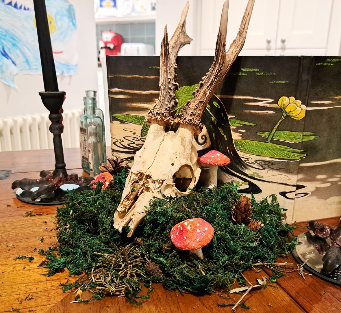 A deer skull surrounded by moss sits in front of a GM screen. There are paper mache fly agaric mushrooms, pine cones, antique bottles and bird leg candlesticks covered in wax drips